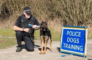 A police officer kneels beside a German Shepherd on a leash. A blue sign reads 