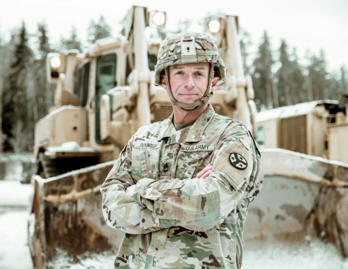 Military engineer in uniform standing confidently beside a snow-covered bulldozer.
